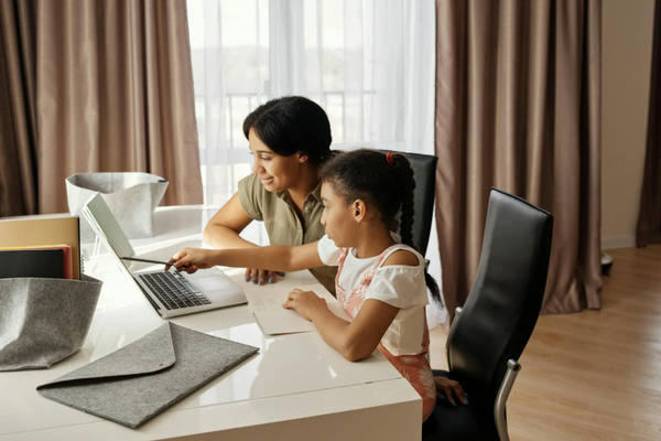 A parent sitting beside a young child at a desk with two laptops