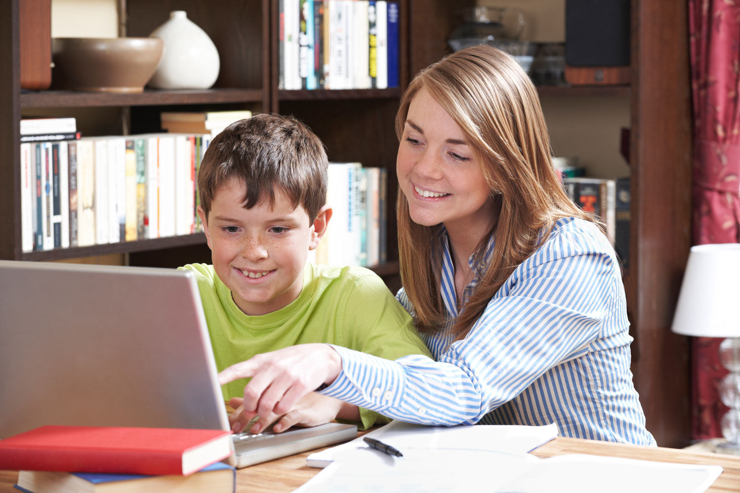 A tutor smiling and guiding a young student working on a laptop, surrounded by books and stationery in a home learning environment. Represents one-on-one online tutoring for personalised education.